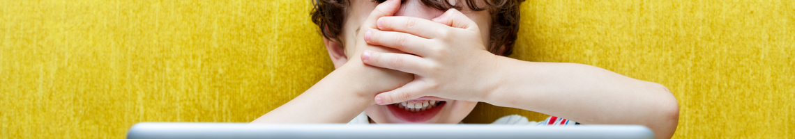 Yellow background. Young child in front of a laptop with their hands over their face